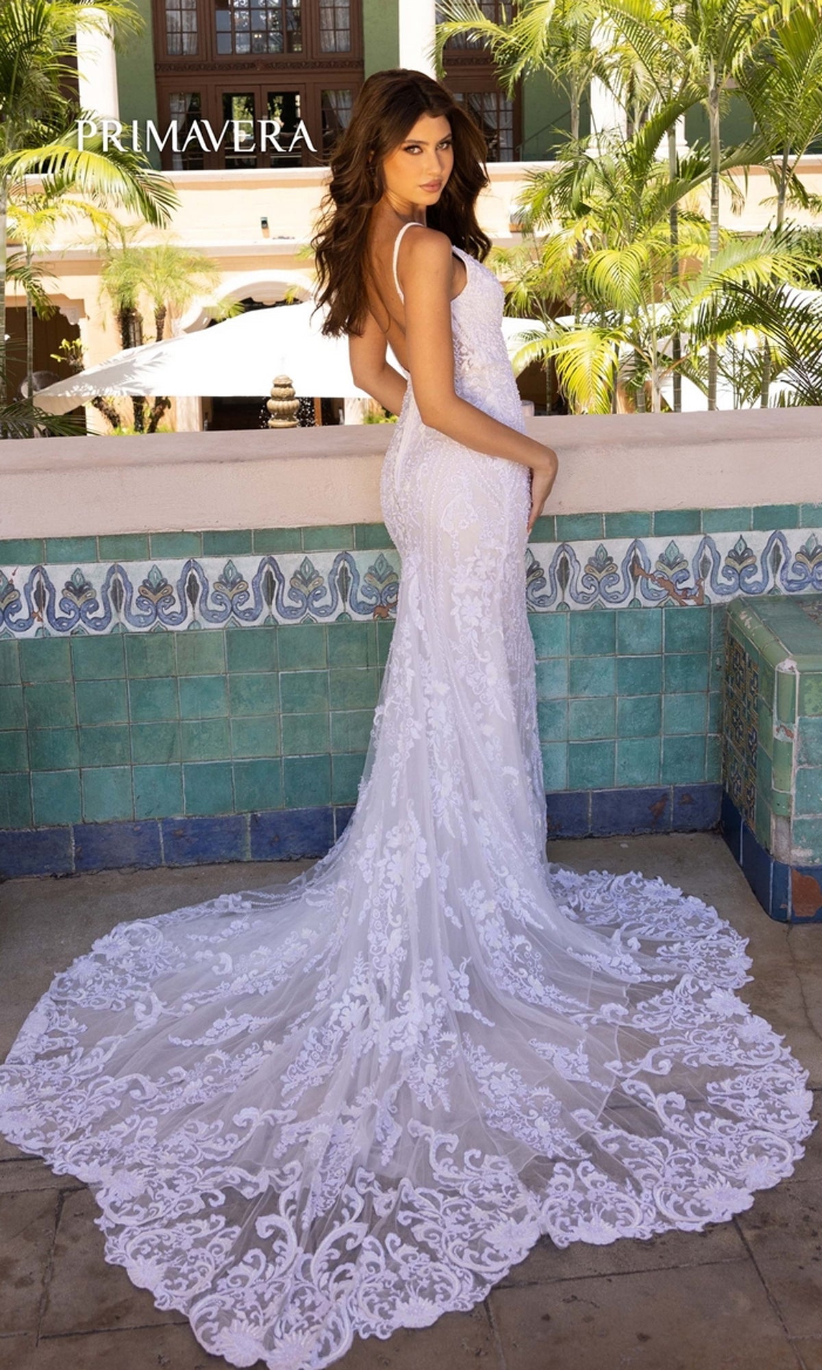 A woman wears the Long Wedding Dress 11106 by Primavera, featuring an ivory lace open back and sweeping train, as she stands on a tiled terrace surrounded by palm trees and lush greenery.
